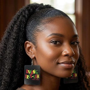 May include: Black square earrings with the text "Living Free-ish Since 1865" in red, yellow, and green. The earrings are hanging from silver hooks. The image shows a person wearing the earrings.