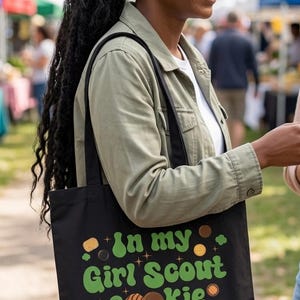 May include: Black tote bag with the text "In my Girl Scout Cookie dealer era" in green and brown. The bag has a design of cookies and stars. The person is wearing a green jacket and white shirt.