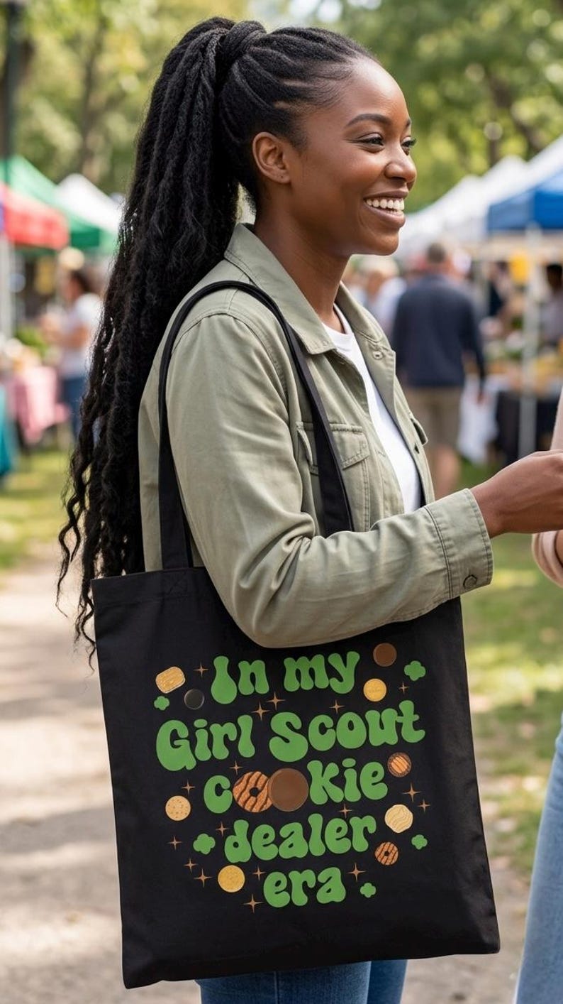 May include: Black tote bag with the text "In my Girl Scout Cookie dealer era" in green and brown. The bag has a design of cookies and stars. The person is wearing a green jacket and white shirt.
