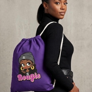 May include: A purple drawstring bag featuring an embroidered cartoon character wearing a gray beanie and gold hoop earrings. The word "Bougie" is written in pink and gold script below the character. The bag has white drawstrings.