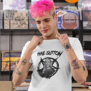 Puede incluir: Una camiseta blanca con un estampado gráfico negro de una mujer en un pentagrama con el texto "Hail Sutton" encima de ella. La persona que lleva la camiseta tiene el pelo rosa y tatuajes en los brazos.
