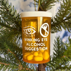 May include: A novelty Christmas tree ornament shaped like a prescription pill bottle. The orange plastic bottle has a white lid and contains yellow pills. Text on the bottle reads "Winking Eye Alcohol Suggestion" with eye and drink symbols. The ornament hangs from a gold string.