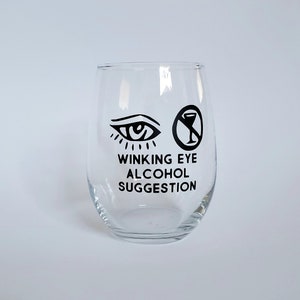 May include: A clear stemless wine glass with black graphics. The graphics include a winking eye and a no-alcohol symbol. Below the graphics is the text "WINKING EYE ALCOHOL SUGGESTION".