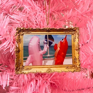 May include: A gold-framed ornament featuring a picture of two inflatable costumes, a pink shark and a red lobster, on a boat. The ornament hangs on a pink Christmas tree.