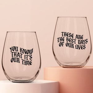 Op de afbeelding: Twee heldere glazen wijnglazen met zwarte tekst. Het glas links zegt "You know that it's our time". Het glas rechts zegt "These are the best days of our lives".