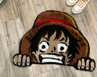 以下が含まれることがあります： 茶色と黒の敷物で、その裏から漫画のキャラクターの顔がのぞいている。キャラクターは黒髪で、麦わら帽子をかぶり、大きく笑っている。