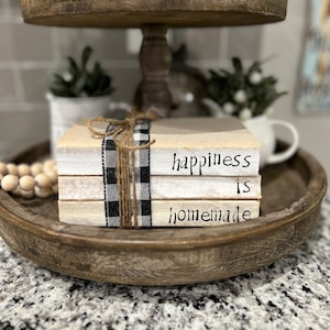 Può includere: Una pila di libri con le parole "happiness is homemade" scritte su di essi. I libri sono avvolti con un nastro a quadri bianco e nero e spago.