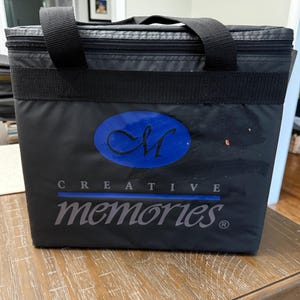 May include: A black rectangular insulated cooler bag with a zippered top and two black nylon handles. The front features a blue oval logo with the letters CM and the text CREATIVE memories printed in white and grey with a blue horizontal line.