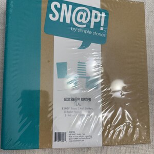 May include: A teal 6x8 inch SN@P! binder by Simple Stories. The binder is sealed in clear plastic and has a brown cover with a teal speech bubble logo. The label indicates it includes pages, dividers, and pocket pages.