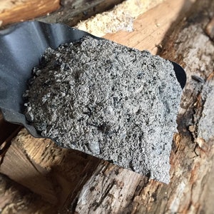 May include: A black metal scoop filled with gray ash, resting on a pile of stacked firewood.
