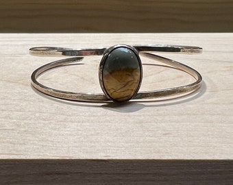 May include: A silver cuff bracelet with an oval, earth-toned stone at the center. The stone features shades of brown, tan, and green. The bracelet has a simple, open design with two parallel bands.