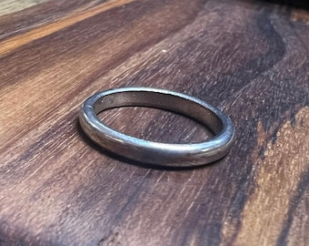 May include: A silver ring with a rounded band. The ring is set against a wooden surface with visible grain patterns. The ring's surface reflects light, highlighting its smooth texture. A simple, classic design.