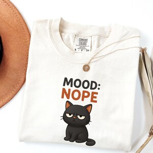 May include: Cream-colored t-shirt with the text "MOOD: NOPE" and a grumpy black cat graphic. A gold necklace and a brown hat are also visible. The shirt is made by Comfort Colors.