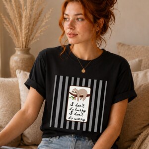 May include: Black t-shirt featuring a sloth illustration on a branch, with the text "I don't do hurry, I don't do worry". The design is set against a background of vertical grey and black stripes.