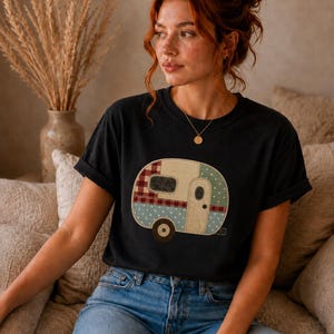 May include: Black t-shirt featuring a colorful camper design. The camper has a red and white plaid window, blue polka dot accents, and a beige door. The shirt is paired with blue jeans.