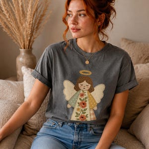 May include: Dark gray t-shirt featuring a distressed angel graphic with a halo and wings. The angel's dress is a patchwork design with floral and polka dot patterns. The shirt is worn with blue jeans.