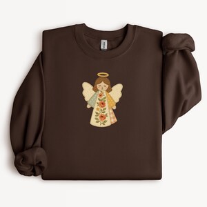 May include: A dark brown sweatshirt featuring a colorful angel graphic. The angel has a gold halo, brown hair, and wings with a patchwork design. The angel's dress has floral and plaid patterns.