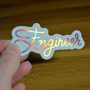 Könnte beinhalten: Holografischer Aufkleber mit dem Wort "Engineer" in Schreibschrift. Der Aufkleber hat einen transparenten Hintergrund und schillernde Buchstaben, die mehrere Farben reflektieren. Der Aufkleber wird vor einer Holzoberfläche gehalten.