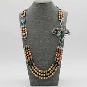 May include: A multi-strand necklace with peach-coloured pearls and silver-toned accents. The necklace has a large pendant with a ram's head design and an abalone shell inlay. The necklace is displayed on a grey mannequin.