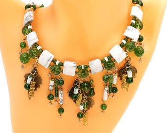 May include: A handmade necklace with white marble-effect square beads, green glass beads, and brass leaf charms. The necklace has a gold chain and is displayed on a mannequin. The design incorporates various shades of green and white.