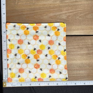 May include: A square fabric cloth featuring a repeating pattern of black bees on a white background with yellow and orange hexagonal honeycomb shapes. A white measuring tape shows the cloth is approximately 7 inches by 7 inches.
