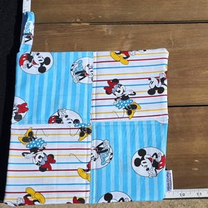 May include: A square fabric potholder featuring a patchwork design of light blue stripes and white fabric with red and yellow striped accents. The fabric is printed with Mickey and Minnie Mouse characters. A white measuring tape shows a length of 28 cm.