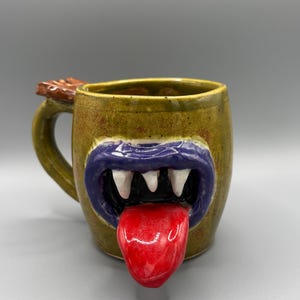 May include: A ceramic mug with a speckled olive green glaze featuring a sculpted monster face. The design includes thick purple lips, sharp white teeth, and a protruding glossy red tongue. A brown textured detail sits atop the handle.