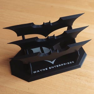 May include: A black Batman logo display stand. The stand features three bat-shaped emblems stacked on top of each other. The base of the stand has the text "WAYNE ENTERPRISES" in white. The stand is made of a matte black material.
