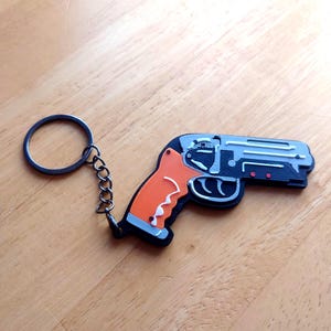 May include: A gun-shaped keyring with a black, grey, and orange design. The keyring has a metal ring and chain. The handle is orange with a white accent, and the gun's body is grey and black.