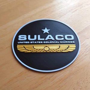 Puede incluir: Un emblema redondo negro con la palabra "SULACO" en blanco, una estrella blanca y alas doradas. El texto "UNITED STATES COLONIAL MARINES" está debajo de la palabra "SULACO". El emblema tiene un borde blanco.