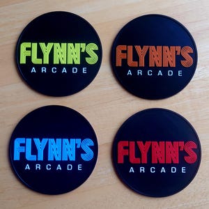 Könnte beinhalten: Vier schwarze, runde Untersetzer mit dem Text "FLYNN'S ARCADE" in verschiedenen Farben: Gelb und Grün, Orange, Blau und Rot. Die Untersetzer sind wahrscheinlich aus einem strapazierfähigen Material.