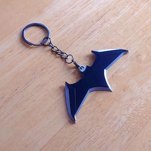 May include: A black metal keyring featuring a bat-shaped pendant with silver-toned beveled edges. The pendant is attached to a circular split ring by a short metal chain. The design is symmetrical with pointed wing tips and a central notch.