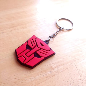 Puede incluir: Un llavero rojo y negro con el símbolo de los Autobots. El llavero tiene un anillo y una cadena de metal plateado. El símbolo de los Autobots es un emblema estilizado con fondo rojo y detalles negros. El llavero es de goma.