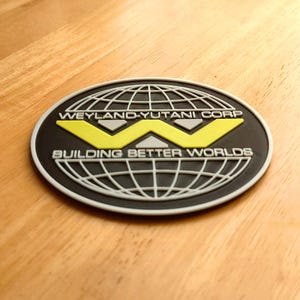May include: An oval-shaped patch with the text "WEYLAND-YUTANI CORP" and "BUILDING BETTER WORLDS". The design features a yellow "W" over a globe graphic, all set against a black background with a white border. The patch is likely an emblem or logo.