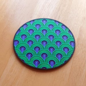 May include: A circular coaster featuring a repeating geometric pattern of concentric semicircles in vibrant green and purple. The design is framed by a solid black border, creating a textured, layered appearance across the entire surface.