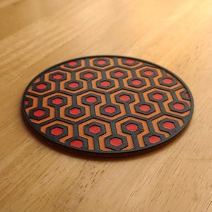 Può includere: Un sottobicchiere rotondo con un motivo geometrico ripetuto in nero, arancione e rosso. Il design presenta una serie di esagoni interconnessi, creando un motivo visivamente sorprendente e simmetrico. Il sottobicchiere è probabilmente realizzato in un materiale resistente.