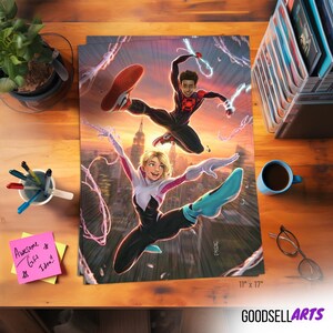 Op de afbeelding: Een kleurrijke poster met twee stripfiguren in actieposities tegen een stadsachtergrond. De poster is 28 x 43 cm groot en bevat de tekst "GOODSELLARTS". Een kopje koffie, een bril en pennen liggen op het houten oppervlak.