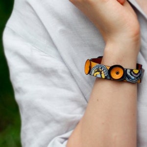 Op de afbeelding: Een kleurrijke armband met gele, blauwe en zwarte cirkels op een bruin leren band.