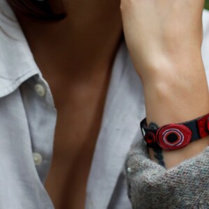 Op de afbeelding: Een close-up van iemands pols met een zwart leren armband met rode en witte ronde versieringen.