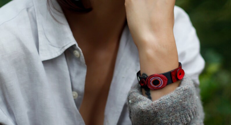 Op de afbeelding: Een close-up van iemands pols met een zwart leren armband met rode en witte ronde versieringen.