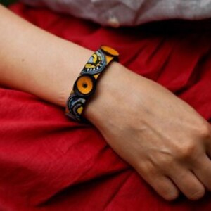 Op de afbeelding: Een stoffen armband met een geel en blauw patroon en houten knopen. De armband wordt om de pols gedragen.