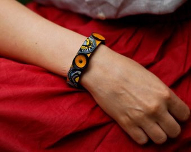 Op de afbeelding: Een stoffen armband met een geel en blauw patroon en houten knopen. De armband wordt om de pols gedragen.