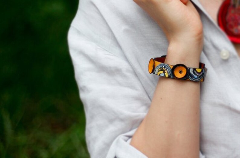Op de afbeelding: Een kleurrijke armband met gele, blauwe en zwarte cirkels op een bruin leren band.