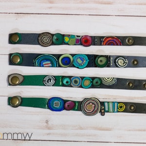 Op de afbeelding: Vier leren armbanden met kleurrijke, gepatroneerde en textuurrijke versieringen. De armbanden zijn genummerd van 1 tot en met 4.