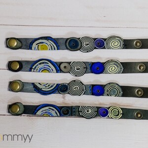Op de afbeelding: Vier grijze leren armbanden met kleurrijke spiraalontwerpen en gouden metalen sluitingen. De armbanden zijn genummerd van 1 tot 4.