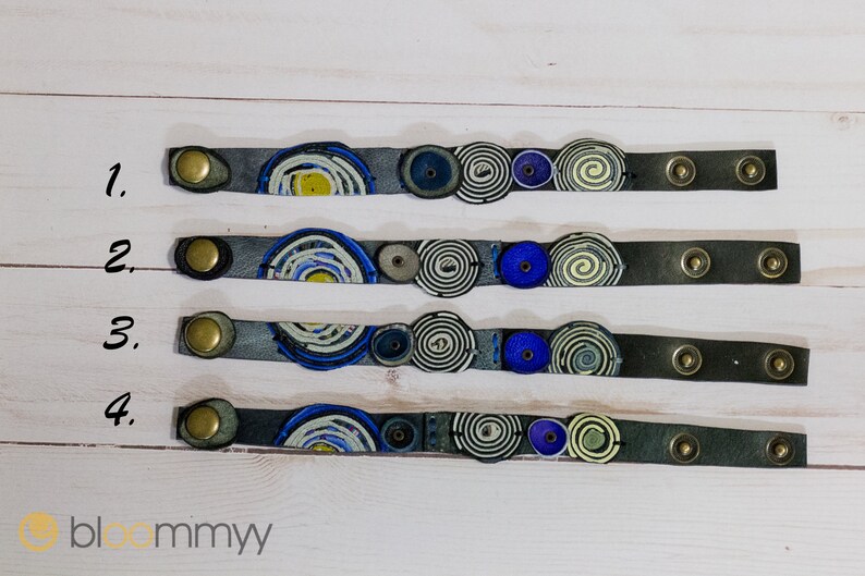 Op de afbeelding: Vier grijze leren armbanden met kleurrijke spiraalontwerpen en gouden metalen sluitingen. De armbanden zijn genummerd van 1 tot 4.