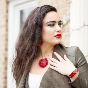 Op de afbeelding: Een vrouw draagt een groene jas met een rode ketting en oorbellen. De ketting en oorbellen zijn gemaakt van stof en hebben een bloemenpatroon.
