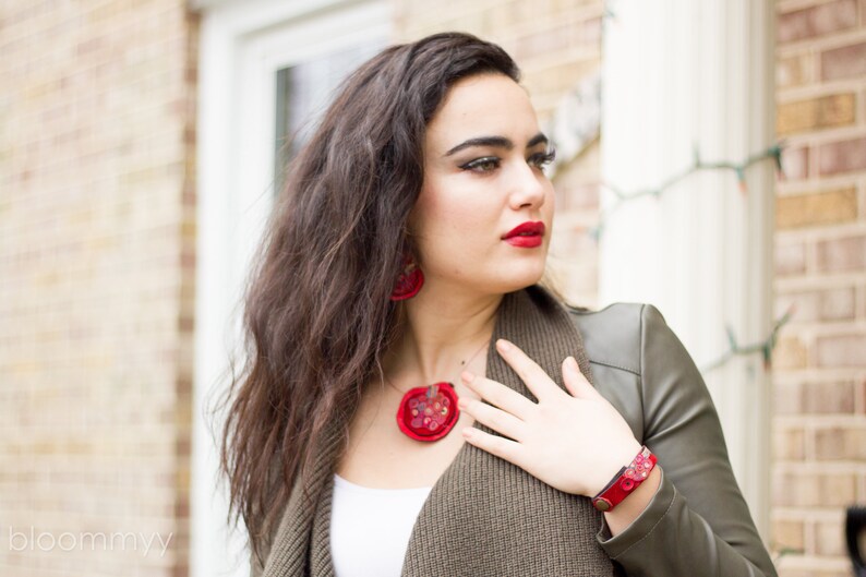 Op de afbeelding: Een vrouw draagt een groene jas met een rode ketting en oorbellen. De ketting en oorbellen zijn gemaakt van stof en hebben een bloemenpatroon.