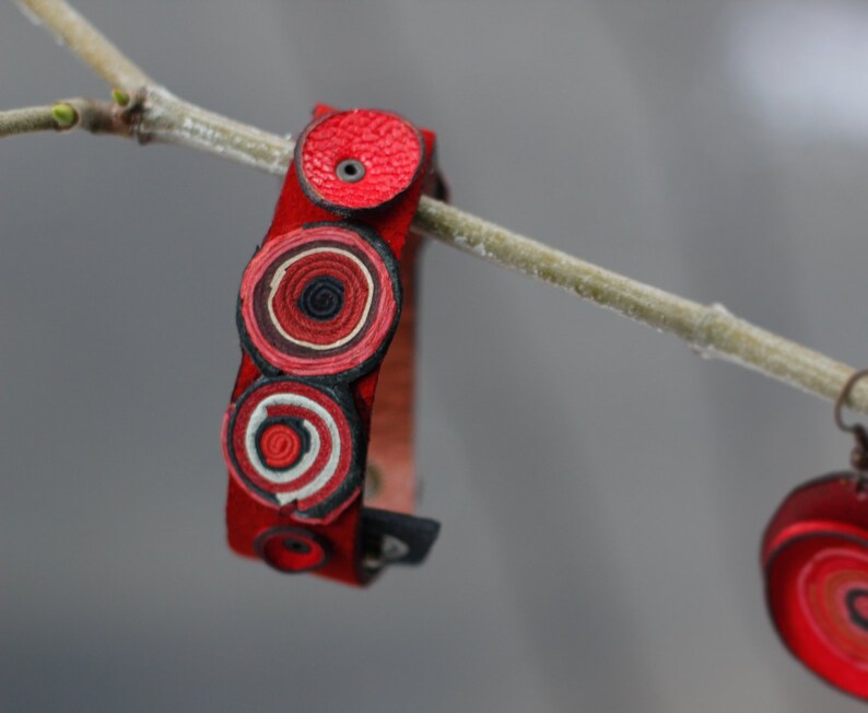 Op de afbeelding: Rood leren armband met drie concentrische cirkels van rood, wit en zwart leer. De armband hangt aan een tak.
