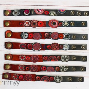 Op de afbeelding: Zeven leren armbanden met rode, witte en zwarte ronde ontwerpen. De armbanden zijn genummerd van 1 tot en met 7.
